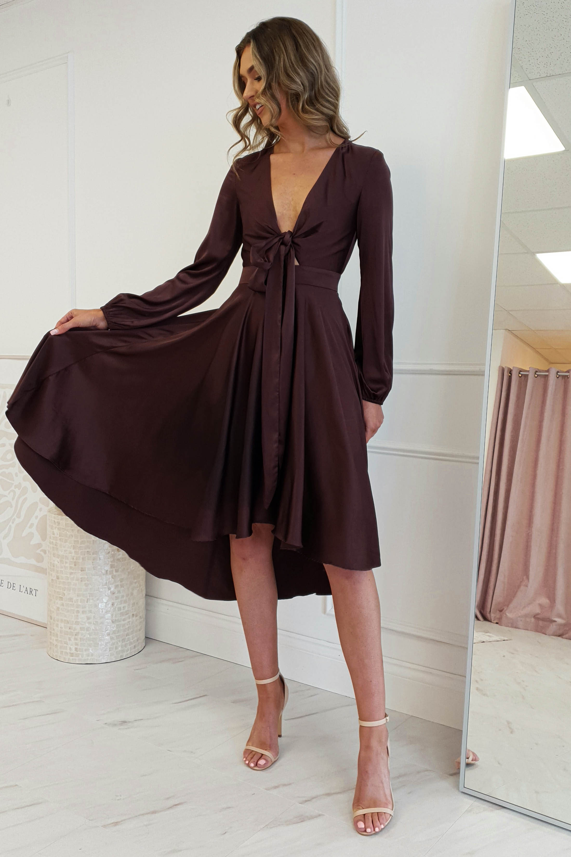 A woman in a stylish, maroon long-sleeve midi dress stands gracefully in a well-lit room. The flowy design features a deep V-neck and bow-tie detail, complemented by her strappy beige heels. She smiles warmly, framed by a curtain and textured pot in the background.