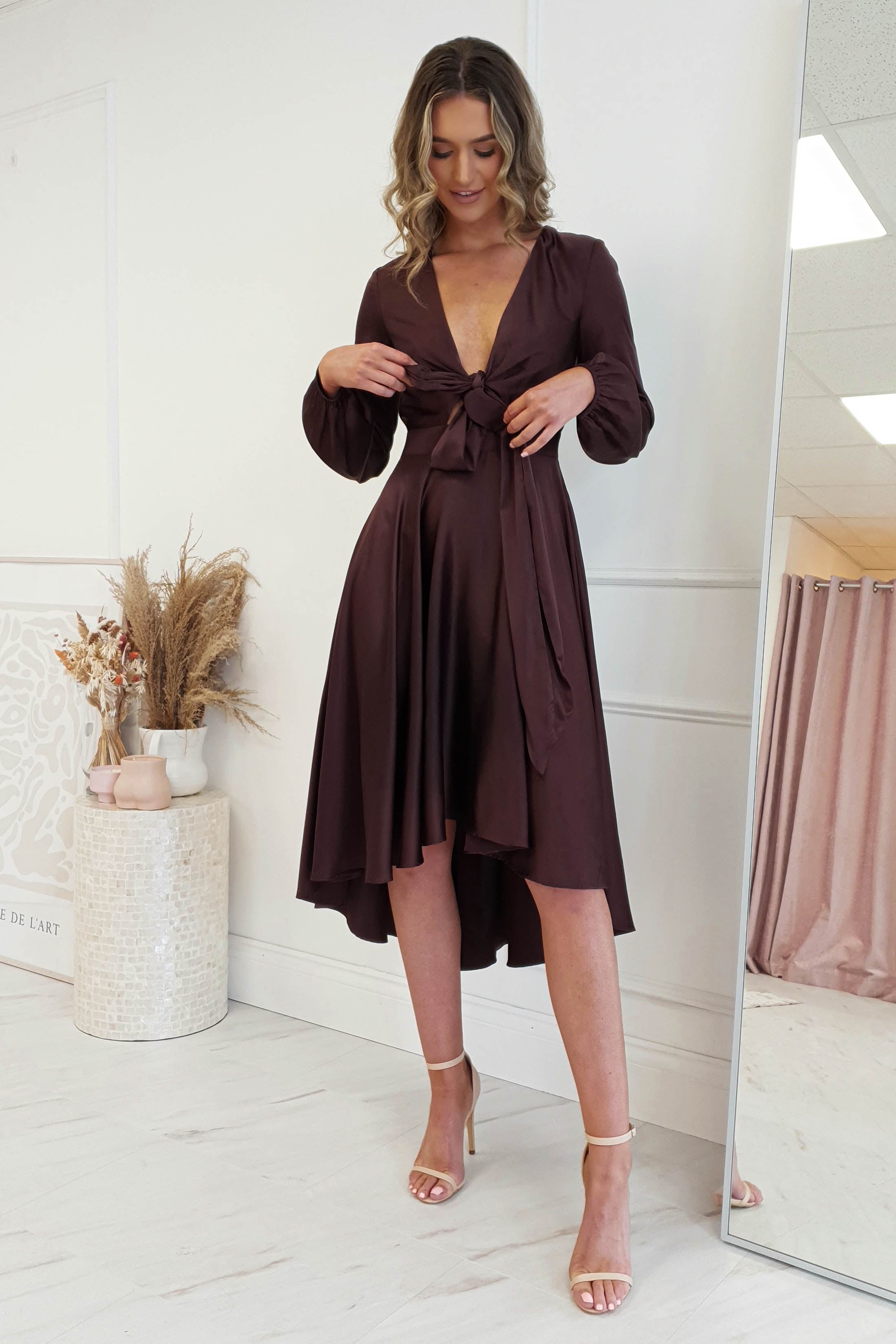 A woman in a stylish, dark brown long-sleeve midi dress with a V-neck and knot design stands indoors. She is wearing beige high heels and smiling while looking down. The room features a vase of dried plants and a large mirror, enhancing the elegant atmosphere.