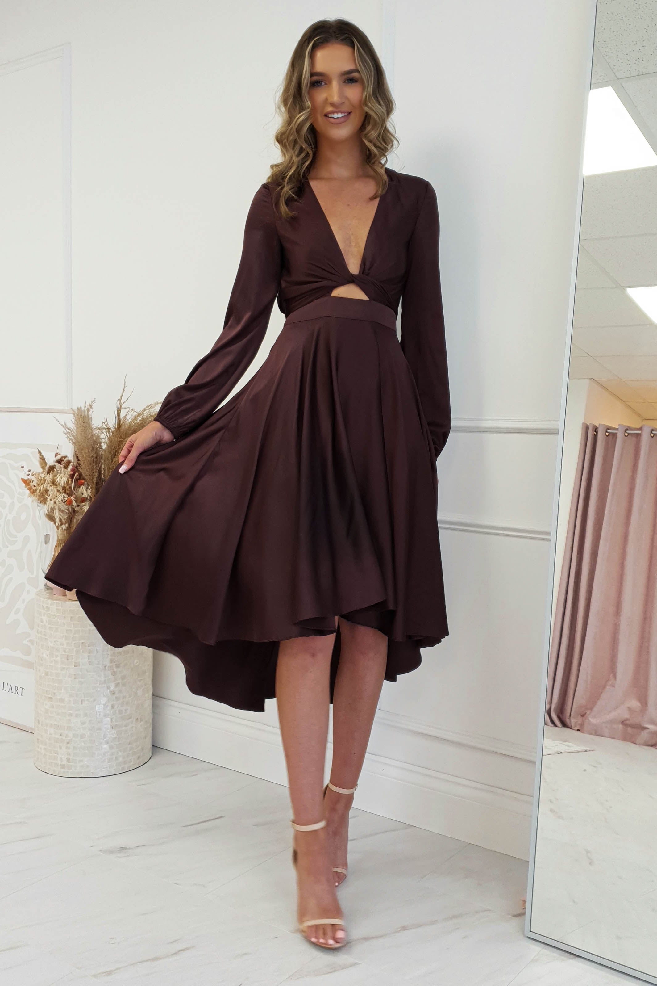A woman in a brown V-neck, long-sleeve midi dress with a high-low hem and waist cutout stands in a well-lit room. She has long, wavy hair and wears strappy heels, smiling at the camera. A large mirror and dried plant décor complete the stylish scene in the background.