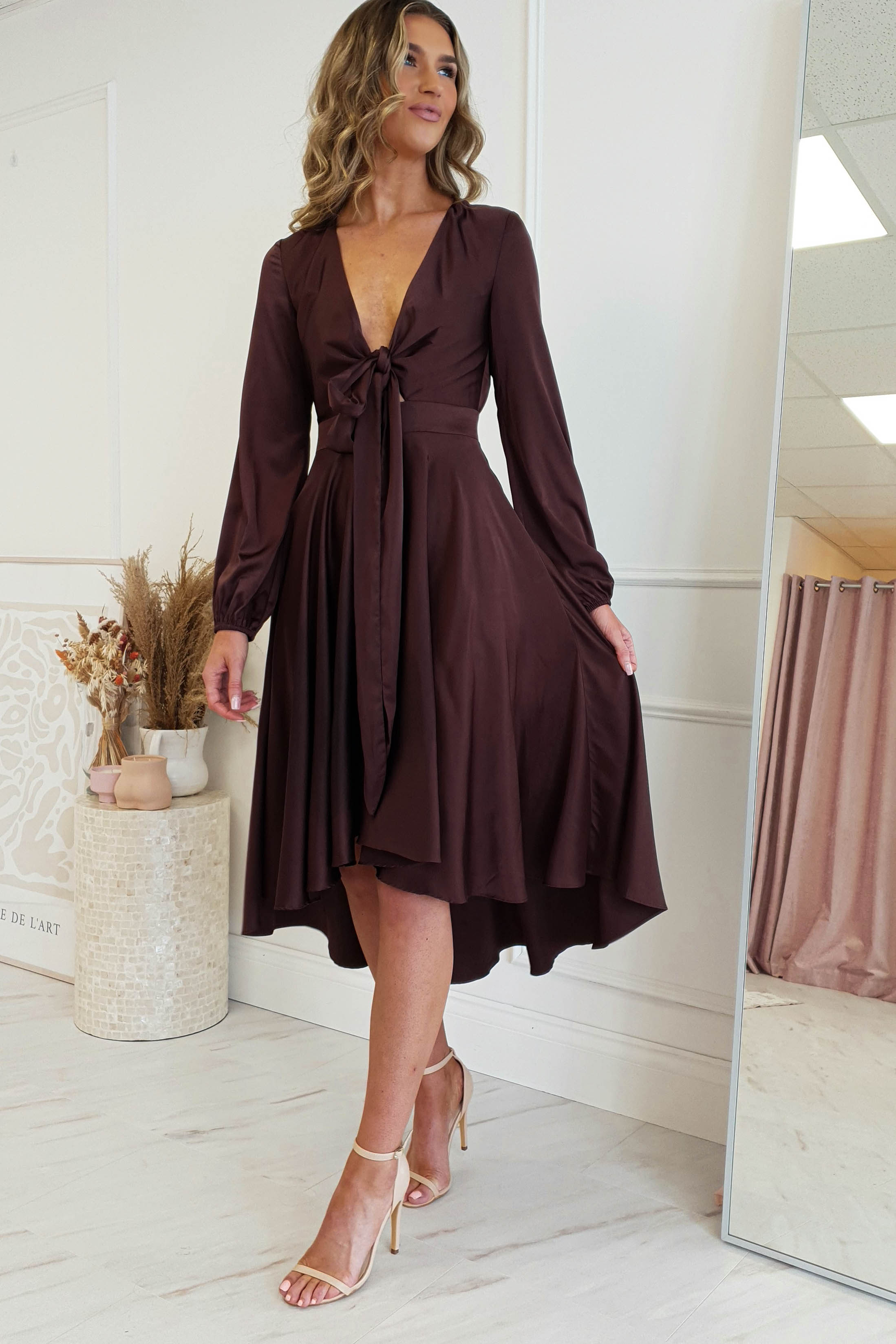 A woman in a deep brown, long-sleeve midi dress with a V-neck and tie knot stands indoors. She is wearing beige high-heeled sandals and is near a large mirror and a decor piece with dried plants. The room has white walls and light-colored flooring, complementing her stylish dress.