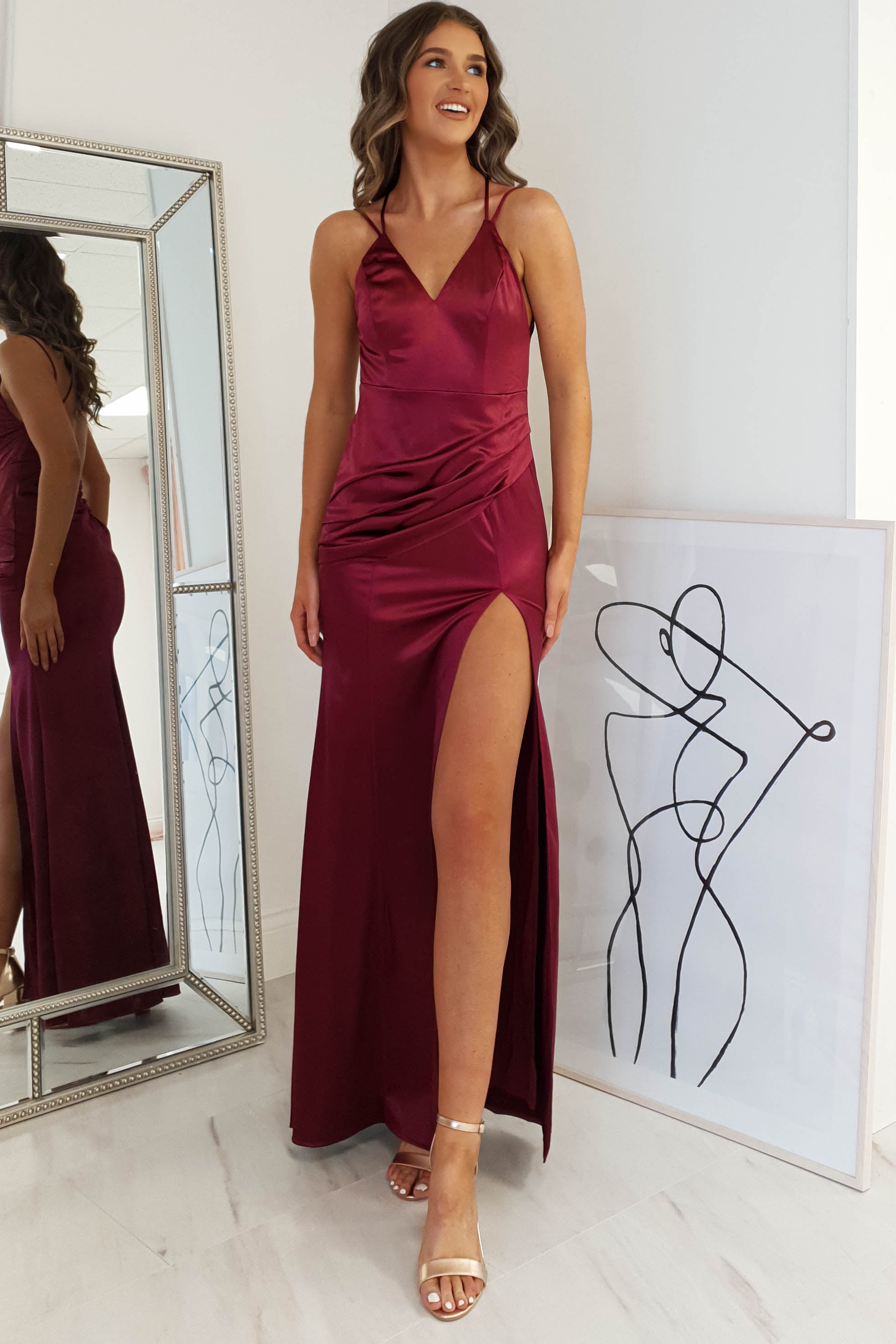 A woman in a sleeveless, deep red satin gown with an open back and high slit stands indoors, smiling. She wears strappy heels, and a framed abstract line drawing rests against the wall beside her. A large mirror reflects her side profile.