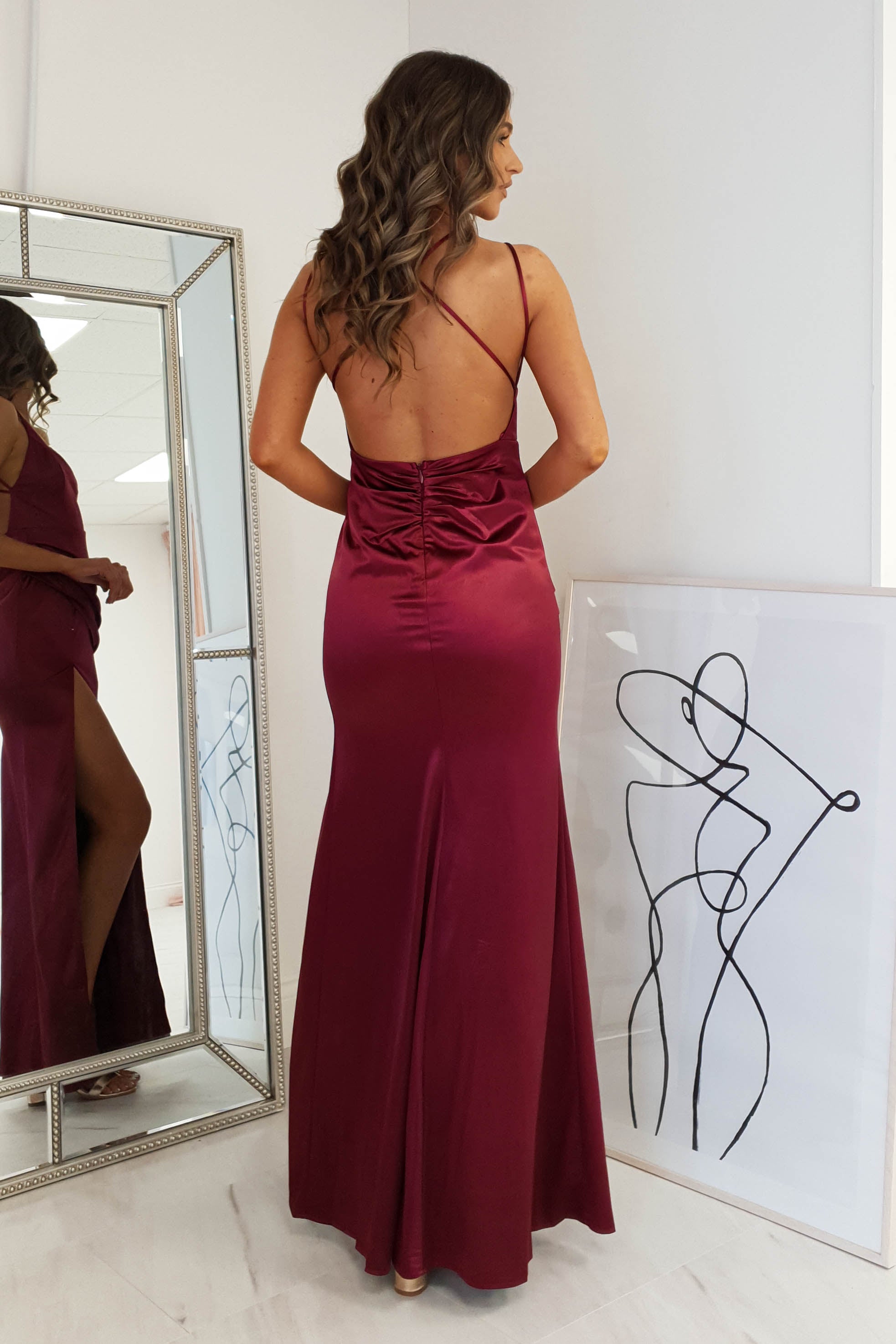 A woman in a floor-length, open back burgundy dress with thin straps stands facing away from the camera. A mirror reflects her side view, and an abstract line art drawing rests against the wall nearby.