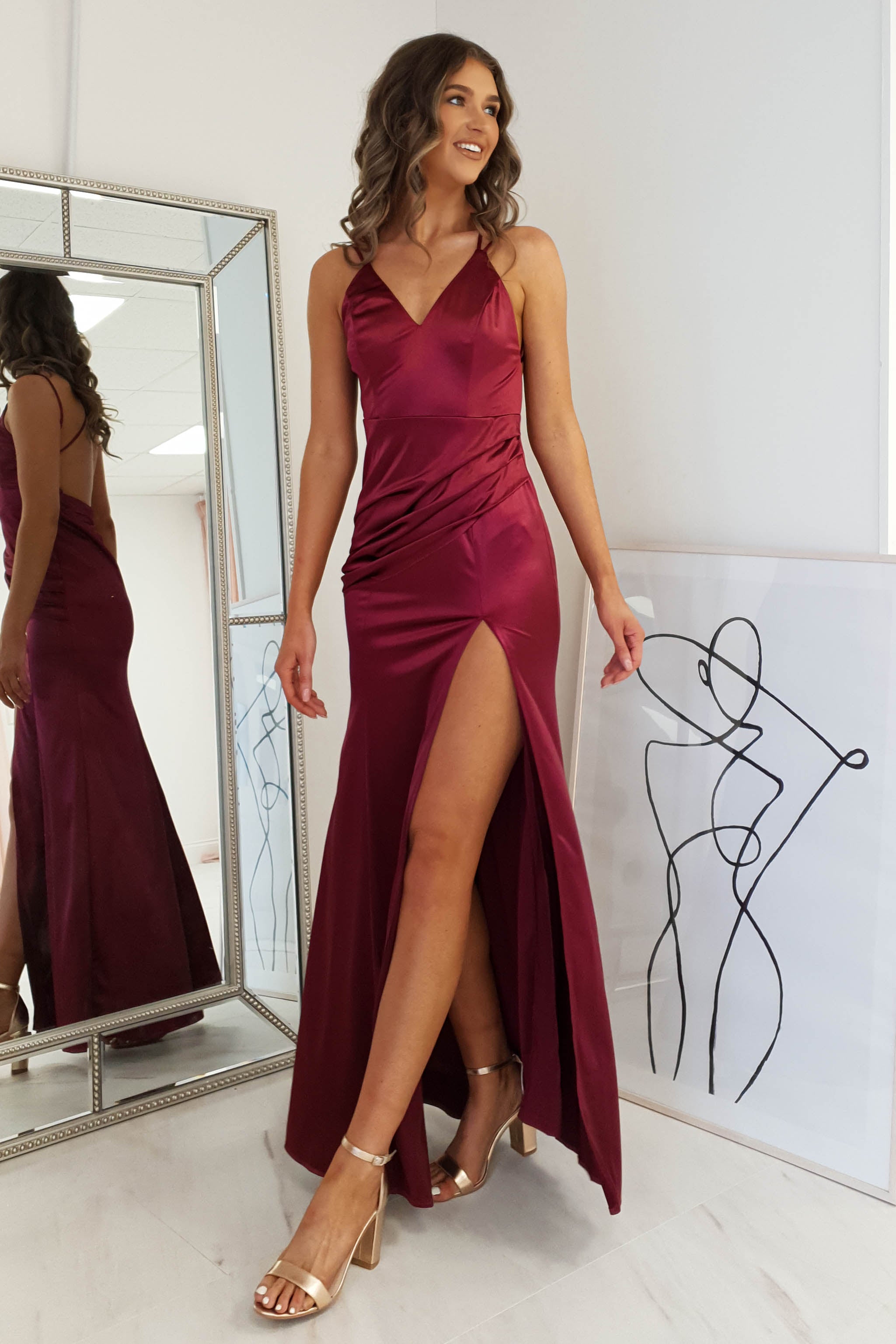 A woman wearing a sleeveless, burgundy dress with a thigh-high slit and open back stands indoors, smiling. She has wavy hair and gold high-heeled sandals. A mirror and minimalist line artwork are visible in the background.
