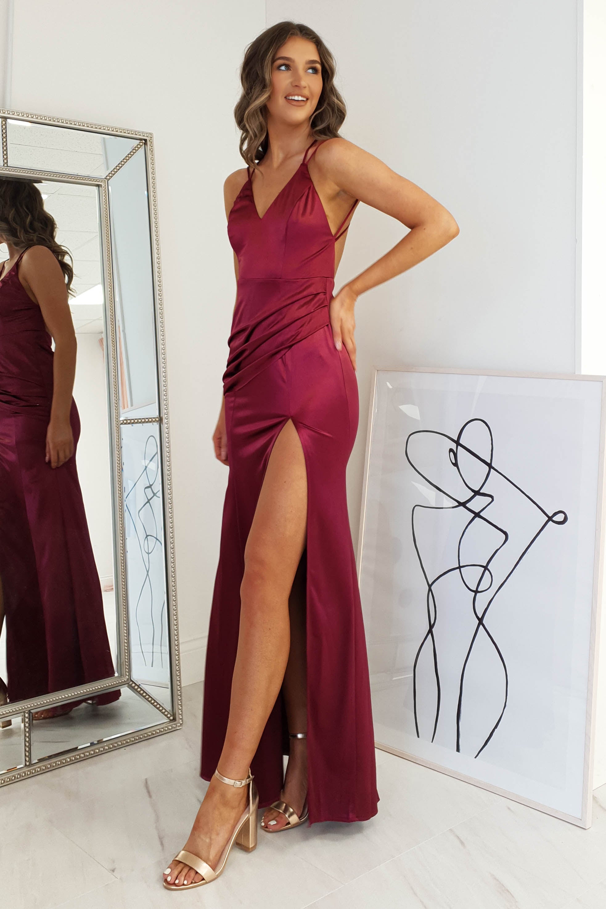 A woman in a burgundy dress with an open back and a thigh-high slit stands smiling beside a framed abstract line drawing and tall mirror, wearing gold high-heeled sandals.