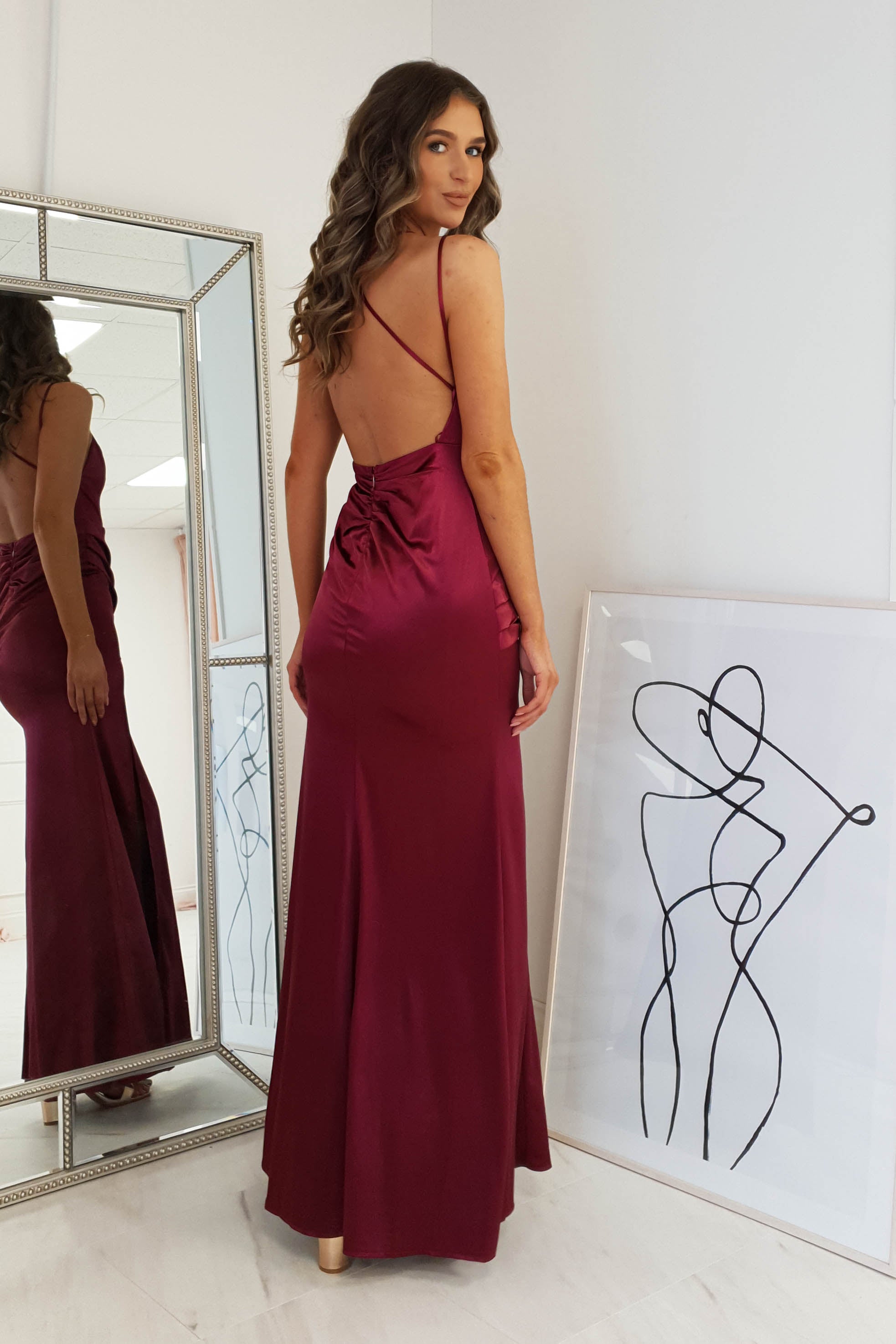 A woman in a burgundy dress with an open back stands before a mirror, her back to the camera. Next to her, a framed abstract line drawing of a figure leans casually against the wall.