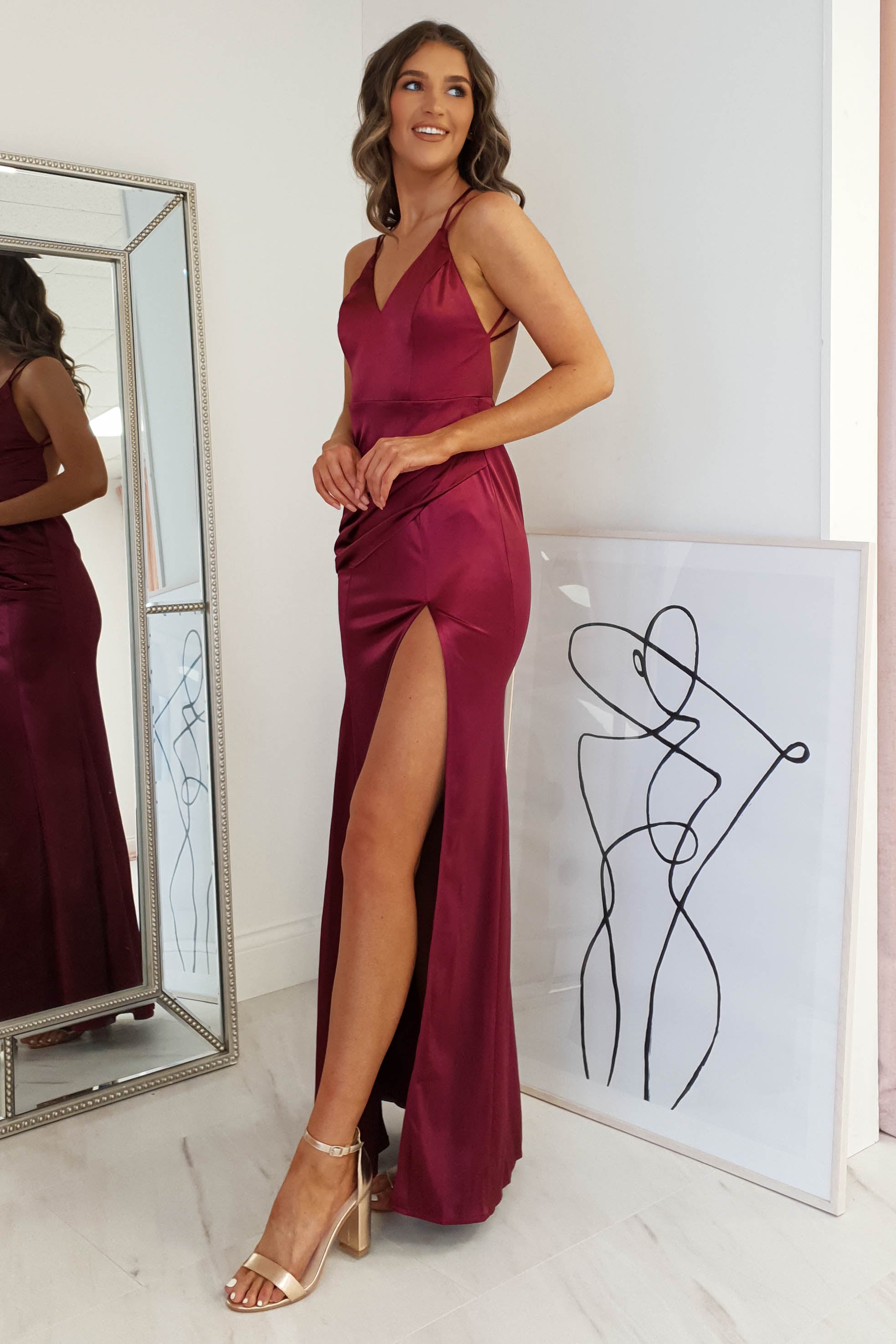 A woman in a sleeveless, burgundy dress with a high slit and open back stands indoors next to a framed abstract line drawing and a large mirror reflecting her side profile. She wears heels and smiles, looking off-camera.
